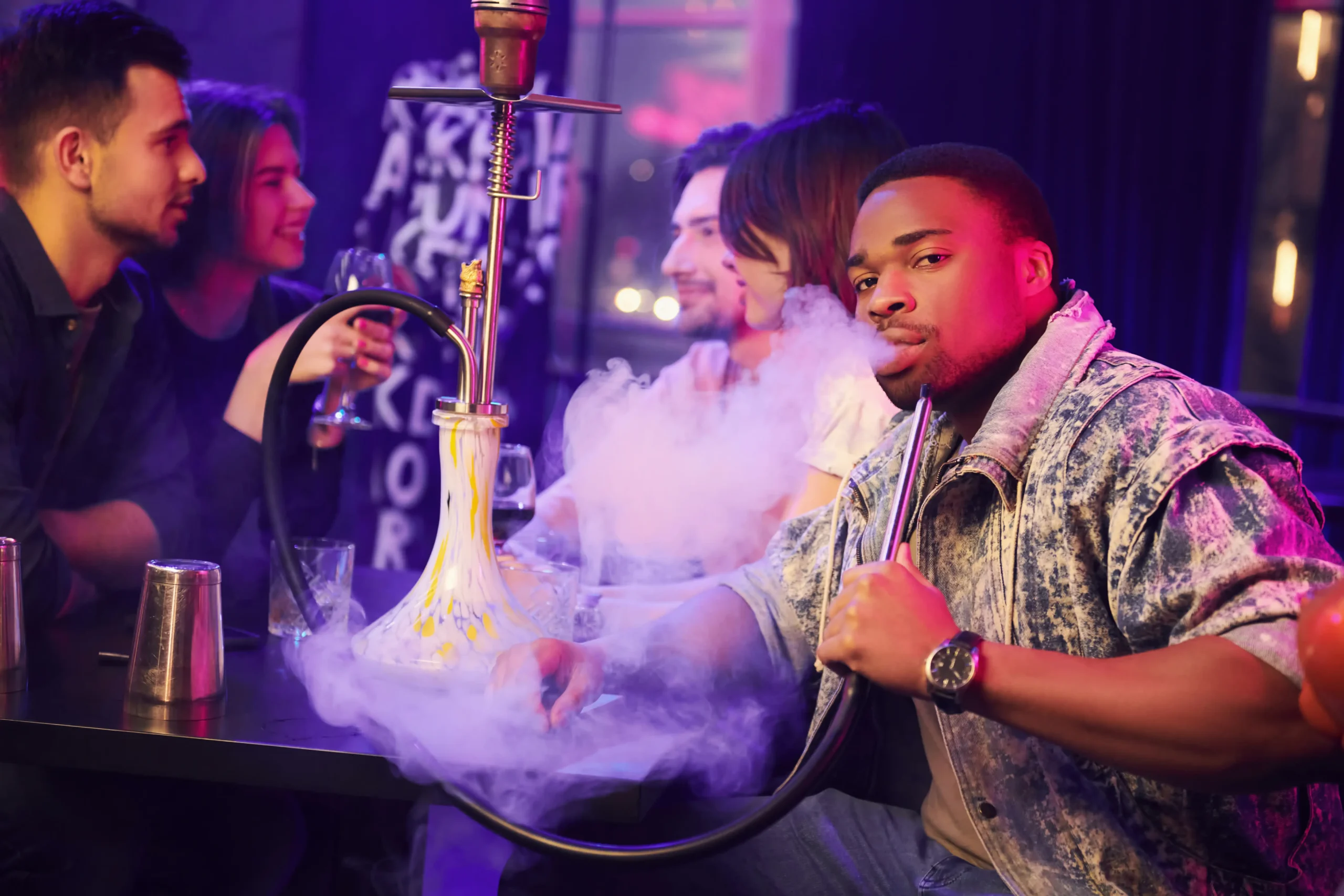 A man smoking and holding a hookah with friends on the table.